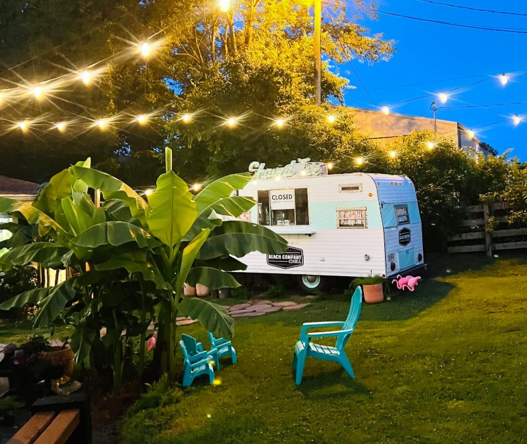 The Beach Co. Chill vintage camper glowing under string lights at dusk, with banana plants and teal Adirondack chairs